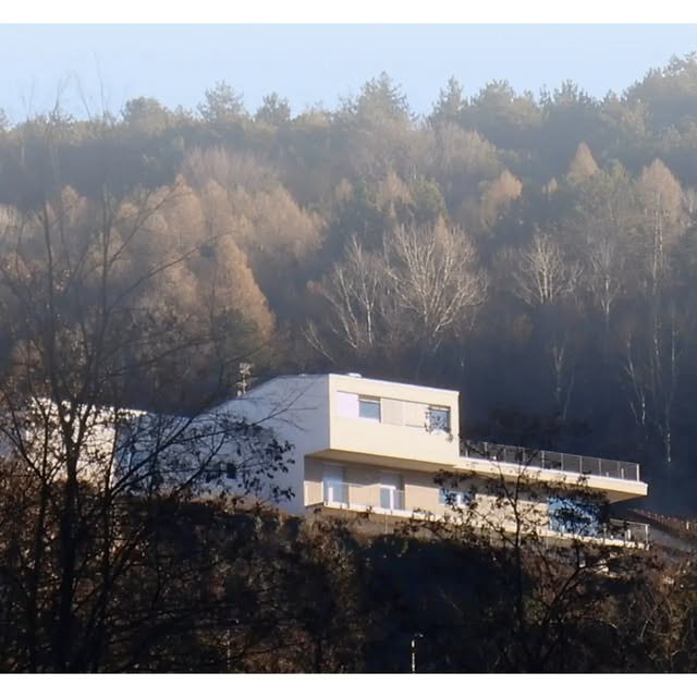 Edificio bianco a volumi sovrapposti su un crinale boscato, con terrazza affacciata a valle
