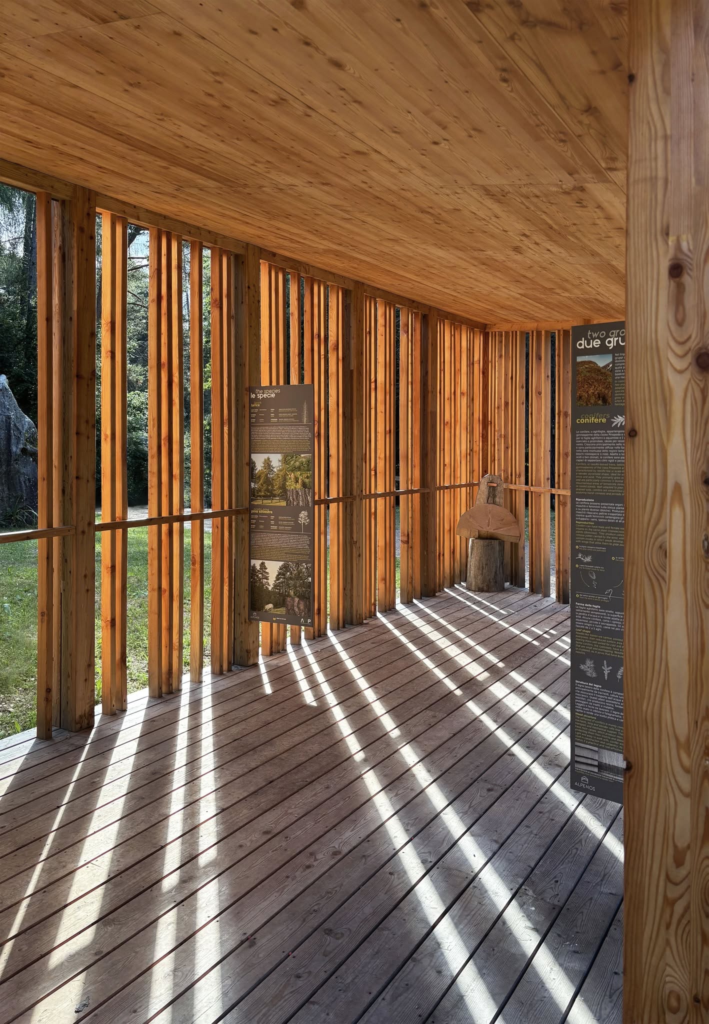 Loggia in legno con montanti verticali e pavimento in tavolato, attraversata dalla luce del pomeriggio